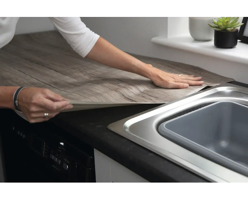 Une femme pose un film décoratif aspect bois sur un plan de travail de cuisine.