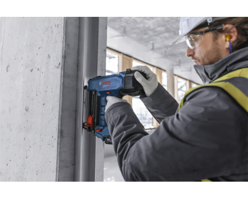 Ein Bauarbeiter befestigt mit einem Bolzensetzgerät ein Metallprofil an einer Betonwand. Bosch Logo.