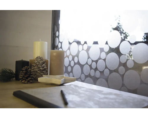 Dekorative Anordnung mit Kerzen, Tannenzapfen, sternförmiger Schale und Notizbuch auf einem Tisch vor einem Fenster mit Punktmusterfolie.