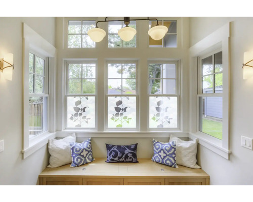 Ein heller Erkerbereich mit Fenstern, dekoriert mit Pflanzenmotiven auf Fensterfolie, Sitzbank mit Kissen, Deckenleuchte und Wandleuchten