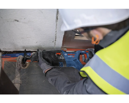 Un ouvrier du bâtiment, coiffé d'un casque de protection et vêtu d'un gilet de sécurité, utilise une scie sabre de marque Bosch pour couper un tuyau.