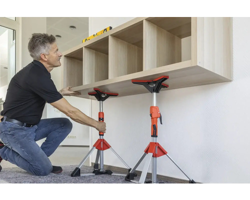 Ein Mann montiert mit Montagestützen ein Holzregal an einer Wand.