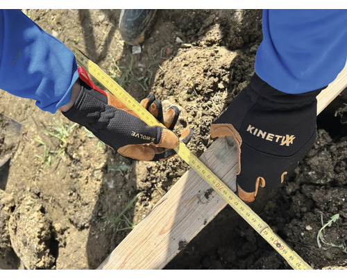 Eine Person misst mit einem Zollstock ein Holzbrett, während sie Arbeitshandschuhe trägt.