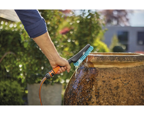 Une personne nettoie un pot avec une brosse raccordée à un tuyau d''arrosage dans le jardin.