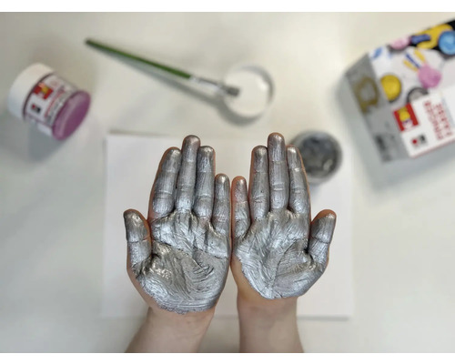 Deux mains recouvertes de peinture argentée, pots de peinture et pinceau sur une table