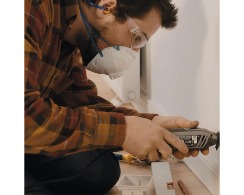 Ein Mann arbeitet mit einem Multifunktionswerkzeug an einer Fussleiste, geschützt mit Atemschutzmaske und Schutzbrille.