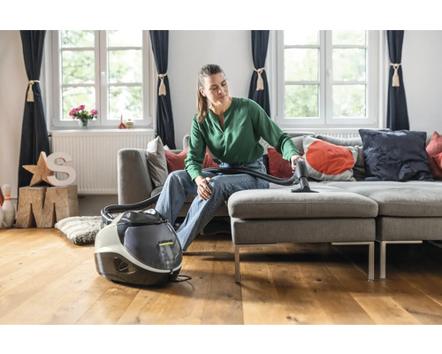 Eine Frau reinigt mit einem Waschsauger ein graues Sofa in einem Wohnzimmer.