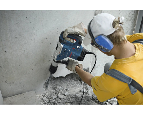 Ein Mann mit Schutzbrille und Gehörschutz bedient einen Abbruchhammer an einer Betonwand.