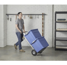 Un homme transporte deux boîtes de rangement bleues avec un diable dans un garage.