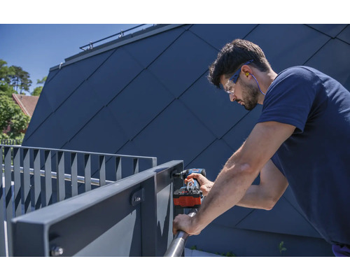 Un homme monte une balustrade métallique avec une visseuse sans fil, portant des lunettes de protection et des protections auditives.