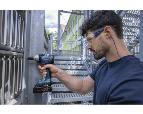Un artisan portant des lunettes de protection et un casque antibruit travaille sur une construction métallique avec une clé à chocs sans fil.