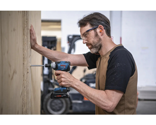 Un homme visse une vis dans une planche de bois à l'aide d'une visseuse à percussion sans fil. Il porte des lunettes de protection et des bouchons d'oreille. Logo Bosch.