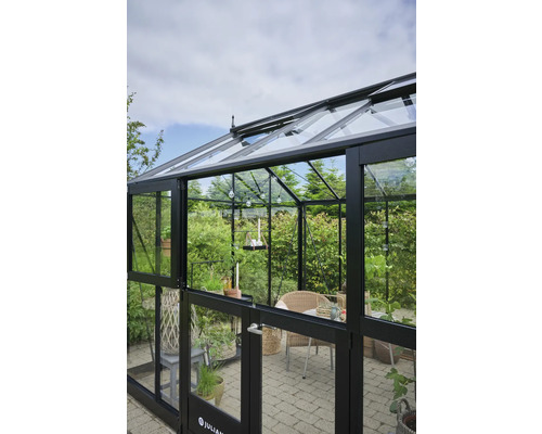 Serre avec parois en verre et cadre noir, équipée de plantes et de sièges.
