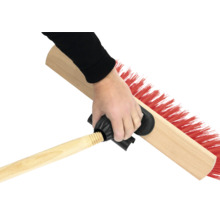 Une main tient une brosse de balayage avec un manche en bois et des poils rouges.