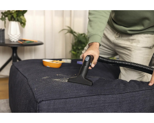 Une personne nettoie un pouf en tissu avec un aspirateur, il y a des miettes dessus.