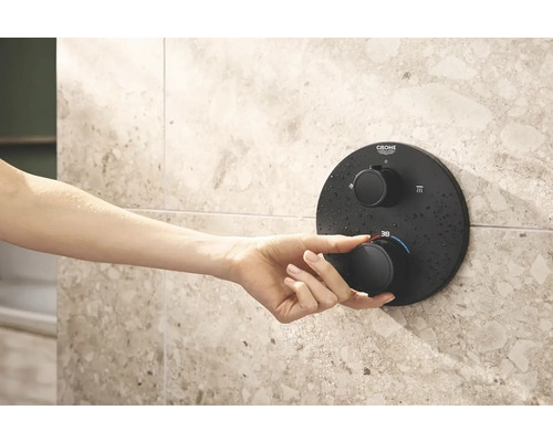 Une main actionne un thermostat de douche Grohe sur un mur carrelé