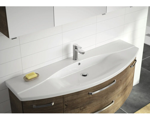 Moderner Waschtisch mit geschwungenem weißem Waschbecken, Einhebelmischer und Unterschrank in Holzoptik vor gefliester Wand.