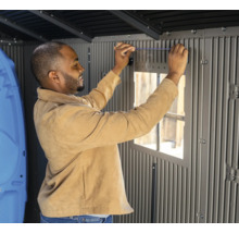 Un homme installe une fenêtre dans une cabane de jardin grise.