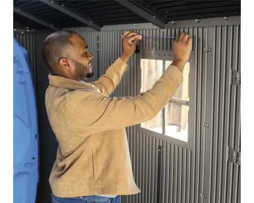 Un homme installe une fenêtre dans une cabane de jardin grise.