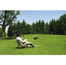 Ein Mann entspannt auf einem Stuhl im Garten und bedient sein Smartphone, während ein Rasenmähroboter das Gras mäht.