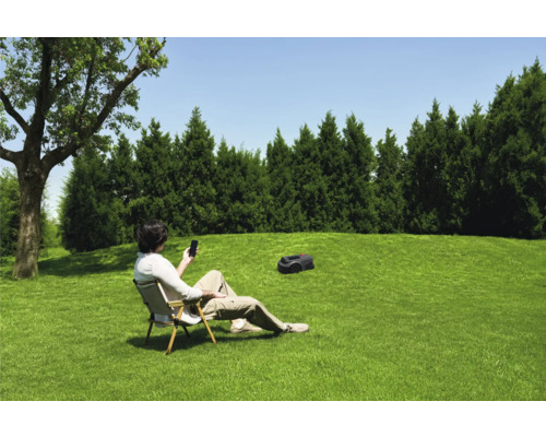 Ein Mann entspannt auf einem Stuhl im Garten und bedient sein Smartphone, während ein Rasenmähroboter das Gras mäht.