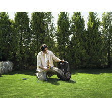 Ein Mann reinigt einen Rasenmähroboter mit einem Gartenschlauch auf einer grünen Rasenfläche vor einer Baumreihe.