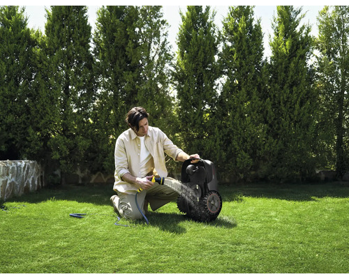 Ein Mann reinigt einen Rasenmähroboter mit einem Gartenschlauch auf einer grünen Rasenfläche vor einer Baumreihe.