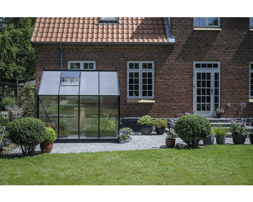Gewächshaus aus Glas und Metall vor einem Backsteingebäude mit Ziegeldach und Rasenfläche.