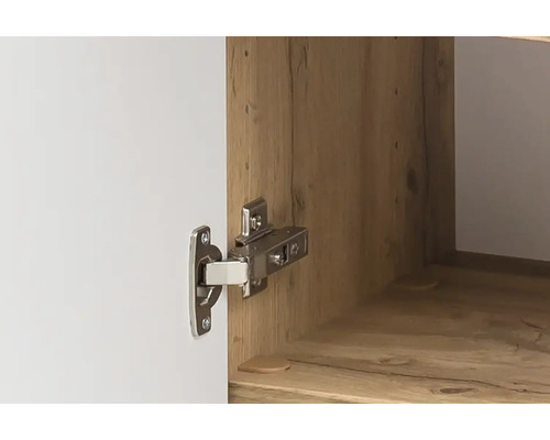 Gros plan d'une charnière de meuble en métal montée sur une porte d'armoire blanche et un intérieur aspect bois.