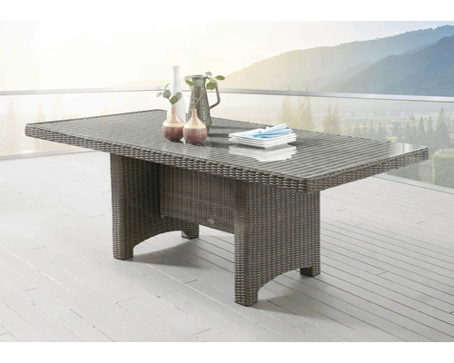 Table de jardin rectangulaire en polyrotin avec plateau en verre sur une terrasse lumineuse devant un paysage de montagne.