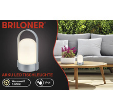 Logo Briloner, lampe de table LED à batterie, blanc chaud 3000 kelvins, IP44, portable avec poignée, dans un jardin sur une table en bois.