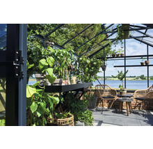 Helles Gewächshaus mit Glaswänden und Metallrahmen, bepflanzt mit diversen Pflanzen sowie Sitzbereich mit Rattansesseln und Blick auf einen See.