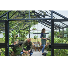 Geräumiges Gewächshaus aus Glas mit schwarzem Rahmen, zwei Personen pflegen Pflanzen in Töpfen, mit Blick auf einen See.