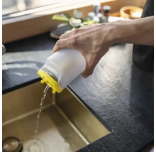 Une personne verse de l'eau d'un récipient en plastique blanc avec un couvercle jaune dans un évier de cuisine doré.
