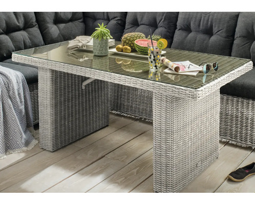 Table de jardin en résine tressée avec plateau en verre devant un salon de jardin avec coussins foncés sur une terrasse.