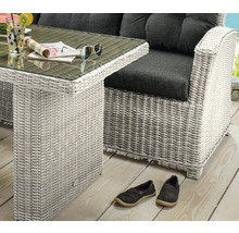 Ensemble de jardin en résine tressée claire avec table en verre et fauteuil lounge aux coussins foncés sur une terrasse en bois.