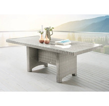 Table de jardin rectangulaire en résine tressée avec plateau en verre sur une terrasse claire devant un paysage de montagne.