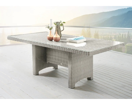 Table de jardin rectangulaire en résine tressée avec plateau en verre sur une terrasse claire devant un paysage de montagne.