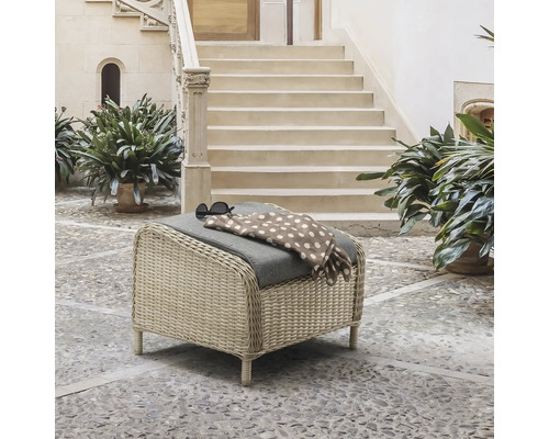 Pouf de jardin en résine tressée avec coussin gris sur une terrasse pavée devant un escalier en pierre.