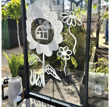 Mit weißer Farbe auf die Glasscheibe eines Gewächshauses gemalte stilisierte Blumen und ein kleines Haus, im Hintergrund Pflanzen.
