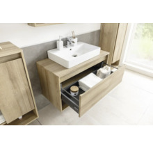 Meuble sous-lavabo moderne à l'aspect bois avec vasque blanche et tiroir ouvert contenant des serviettes et des accessoires.