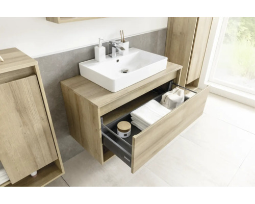 Meuble sous-lavabo moderne à l'aspect bois avec vasque blanche et tiroir ouvert contenant des serviettes et des accessoires.