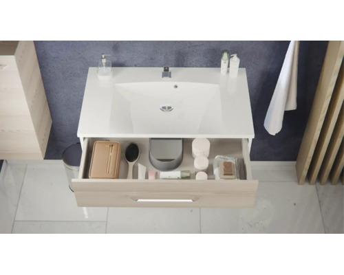 Meuble sous-vasque moderne avec lavabo blanc et tiroir ouvert aspect bois contenant des accessoires de salle de bains.