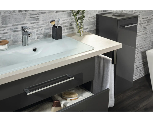 Meuble de lavabo moderne avec vasque en verre, meuble bas anthracite et colonne assortie devant un mur aspect pierre.