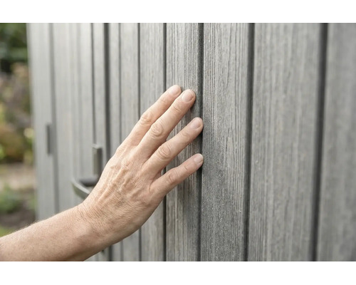 Main touchant un revêtement mural gris aspect bois avec une structure de surface tactile.