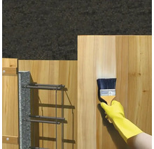 Une main gantée applique un produit de protection pour le bois à l'aide d'un pinceau sur une planche en bois.