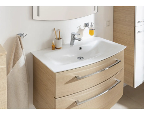 Meuble de salle de bains moderne avec meuble sous-vasque incurvé aspect chêne, vasque blanche, robinet chromé et accessoires.
