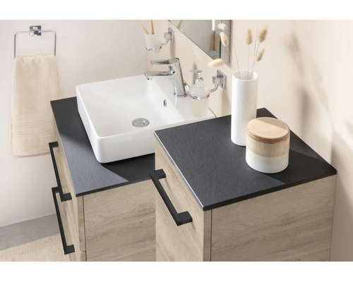 Salle de bain moderne avec vasque à poser blanche, plan de travail sombre et meuble sous-vasque aspect chêne avec poignées noires.