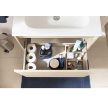 Tiroir ouvert d'un meuble sous-vasque clair avec des accessoires de salle de bains comme du papier toilette et des produits de soin sous un lavabo blanc.