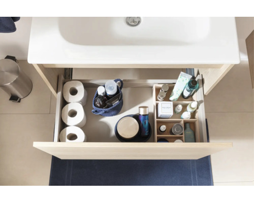 Tiroir ouvert d'un meuble sous-vasque clair avec des accessoires de salle de bains comme du papier toilette et des produits de soin sous un lavabo blanc.